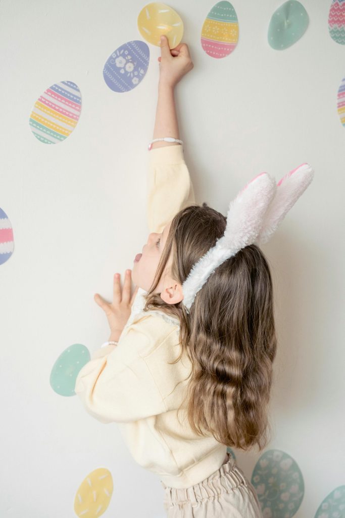 Seitenansicht: Kleines Mädchen mit Hasenohren klebt bunte Osteraufkleber an eine weiße Wand in der Kindertagesstätte.